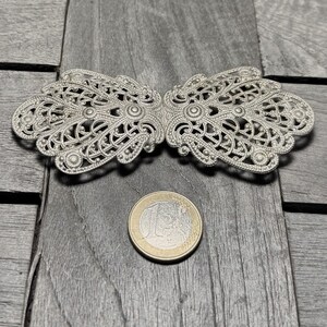 K&ouml;nnte beinhalten: Eine silberne Filigran-Schnalle mit einem zarten, filigranen Design. Die Schnalle ist aus Metall und hat eine gl&auml;nzende Oberfl&auml;che.