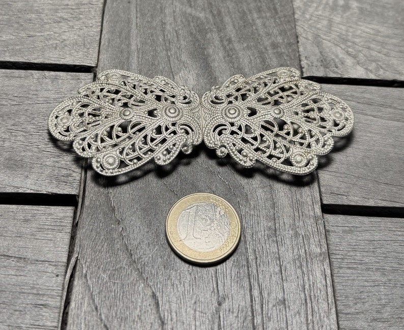 K&ouml;nnte beinhalten: Eine silberne Filigran-Schnalle mit einem zarten, filigranen Design. Die Schnalle ist aus Metall und hat eine gl&auml;nzende Oberfl&auml;che.