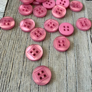 Può includere: Una collezione di bottoni rosa con vari design. Alcuni bottoni hanno quattro fori, mentre altri presentano un design quadrato con quattro fori più piccoli. I bottoni sono sparsi su una superficie di legno, con un tema di artigianato o cucito.