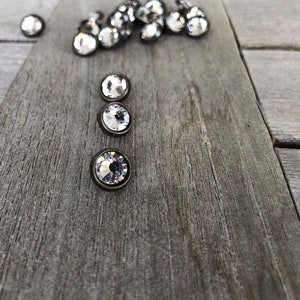 May include: A close-up of three clear crystal buttons with silver settings. The buttons are arranged vertically on a wooden surface.