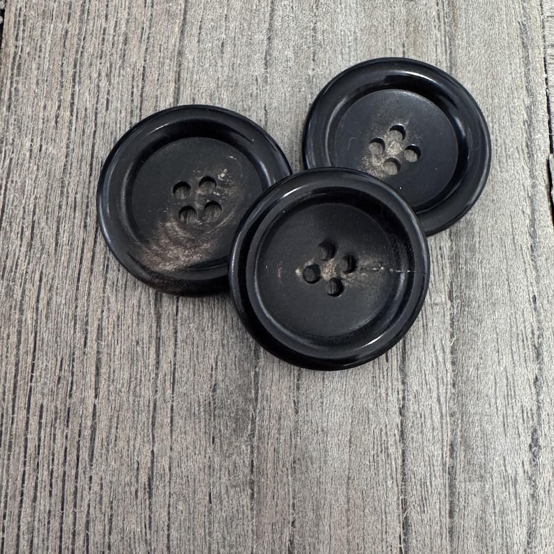 K&ouml;nnte beinhalten: Drei schwarze Kn&ouml;pfe mit jeweils vier L&ouml;chern. Die Kn&ouml;pfe liegen auf einer Holzoberfl&auml;che.