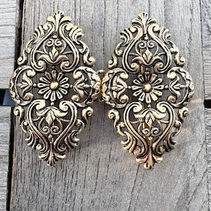 May include: Ornate gold-colored metal clasp with an intricate floral and scroll design. The clasp is composed of two symmetrical halves connected by a hinge. The design includes stylized flowers and swirling patterns, creating a vintage aesthetic.