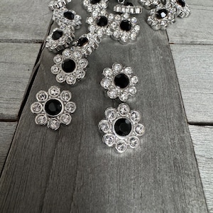 Könnte beinhalten: Eine Sammlung silberfarbener, blumenförmiger Knöpfe mit schwarzen Strasssteinen in der Mitte. Die Knöpfe sind auf einer grauen Holzoberfläche verstreut.