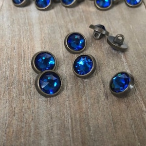 May include: A group of 20 blue rhinestone buttons with antique brass settings. The buttons are arranged in a semi-circle on a wooden surface.