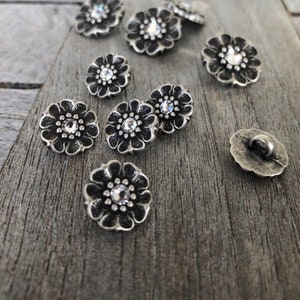 May include: A collection of silver-toned metal flower-shaped buttons with clear rhinestone centres. The buttons are scattered on a wooden surface.