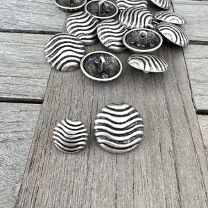 Peut inclure: Un ensemble de boutons argentés avec un motif ondulé. Les boutons sont disposés en pile sur une surface en bois.