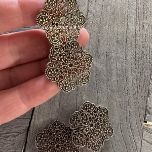 Könnte beinhalten: Drei goldfarbene filigranartige Metallverzierungen in Form einer Blume. Die Verzierungen sind filigran und detailliert, mit einem zarten Design.