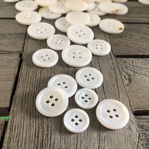 Puede incluir: Una colección de botones de nácar blanco, cada uno con cuatro agujeros. Los botones están dispuestos en una línea sobre una superficie de madera.