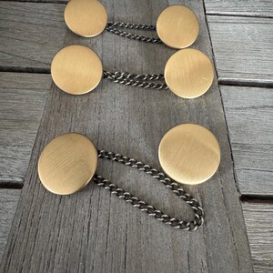Könnte beinhalten: Drei Sätze goldfarbener Manschettenknöpfe aus Metall mit einer Kette, die die beiden Teile verbindet. Die Manschettenknöpfe sind rund und haben eine gebürstete Oberfläche.