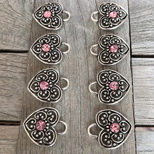 May include: Eight silver heart-shaped charms with pink rhinestone accents. Each charm has a loop for attaching to a bracelet or necklace.