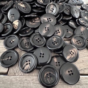 K&ouml;nnte beinhalten: Ein Haufen schwarzer und goldener Kn&ouml;pfe mit jeweils vier L&ouml;chern. Die Kn&ouml;pfe sind aus einem glatten, gl&auml;nzenden Material gefertigt.