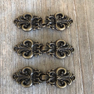 May include: Three antique gold-toned metal hook and eye closures. Each closure features a decorative, ornate design with a scroll-like pattern.