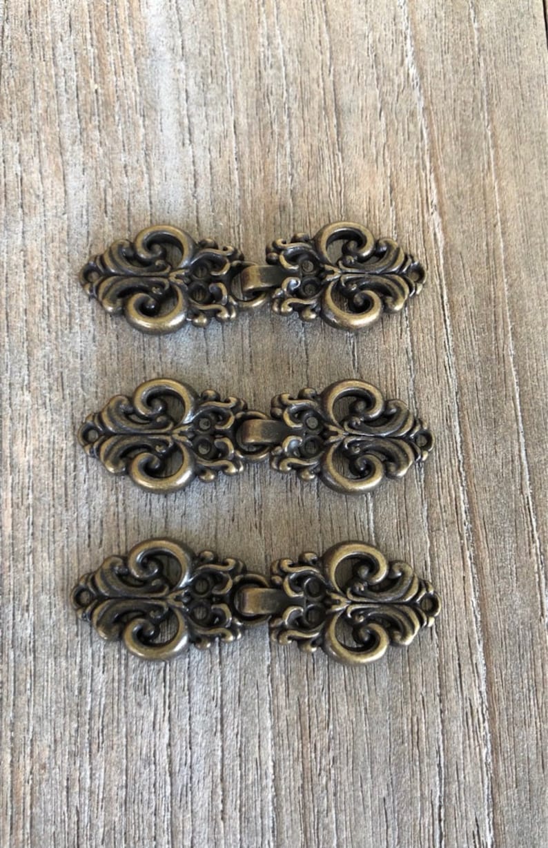 May include: Three antique gold-toned metal hook and eye closures. Each closure features a decorative, ornate design with a scroll-like pattern.