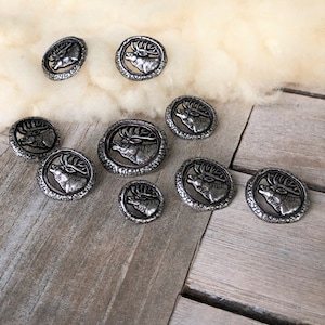 Könnte beinhalten: Ein Satz von acht silberfarbenen Metallknöpfen mit einem erhabenen Hirschkopfdesign. Die Knöpfe sind rund und haben eine strukturierte Oberfläche.