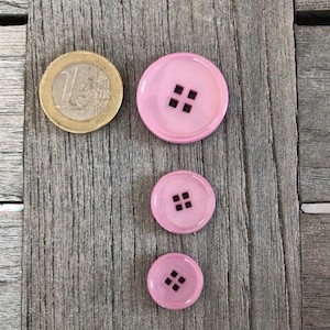 May include: Three pink plastic buttons with four holes each. The buttons are arranged in a vertical line, with the largest button at the top and the smallest button at the bottom.