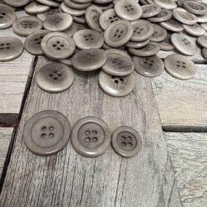 Puede incluir: Un montón de botones de plástico marrón de varios tamaños. Los botones tienen cuatro agujeros cada uno. Tres botones están separados del montón y están dispuestos en una fila de mayor a menor.