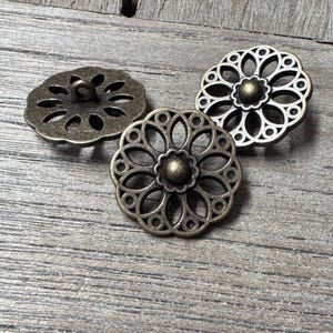 May include: Three antique gold-toned metal buttons with a decorative floral design. The buttons have a raised centre and a loop on the back for sewing.
