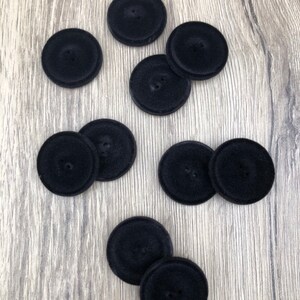 May include: Nine black velvet buttons with two holes each. The buttons are arranged in a scattered pattern on a wood surface.