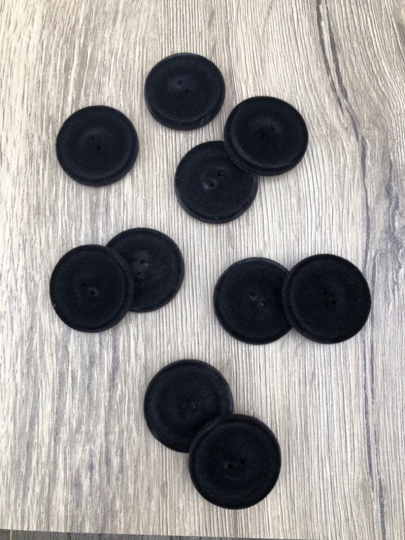 May include: Nine black velvet buttons with two holes each. The buttons are arranged in a scattered pattern on a wood surface.