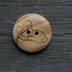 May include: A round wooden button with a carved design of a sleeping dog. The button is light brown with two small holes for sewing. The dog is lying down with its eyes closed.