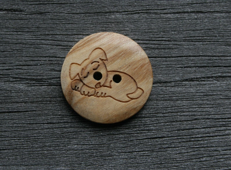May include: A round wooden button with a carved design of a sleeping dog. The button is light brown with two small holes for sewing. The dog is lying down with its eyes closed.