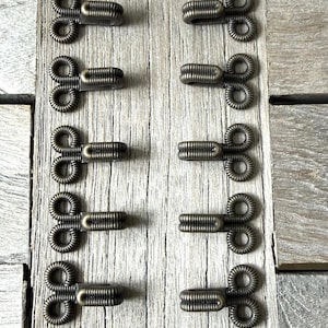 May include: Close-up of antique brass hook and eye closures. The closures are arranged in two columns of five, with the hooks and eyes alternating. The background is a weathered, light-coloured wooden surface.