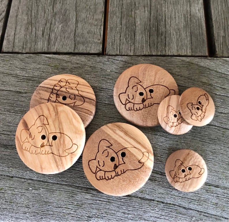 May include: A collection of wooden buttons with a laser-etched design of a sleeping puppy. The buttons vary in size and feature a light brown color with visible wood grain. The puppy design is in a simple, cartoon style.