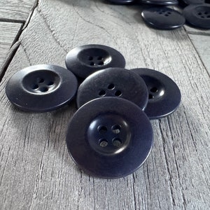 Puede incluir: Un grupo de seis botones de color azul oscuro con cuatro agujeros cada uno. Los botones están dispuestos en semicírculo sobre una superficie de madera.