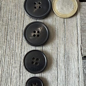 May include: Four black buttons of varying sizes with four holes each, arranged vertically on a wooden surface. A one Euro coin is next to the largest button. The buttons are round and have a glossy finish.