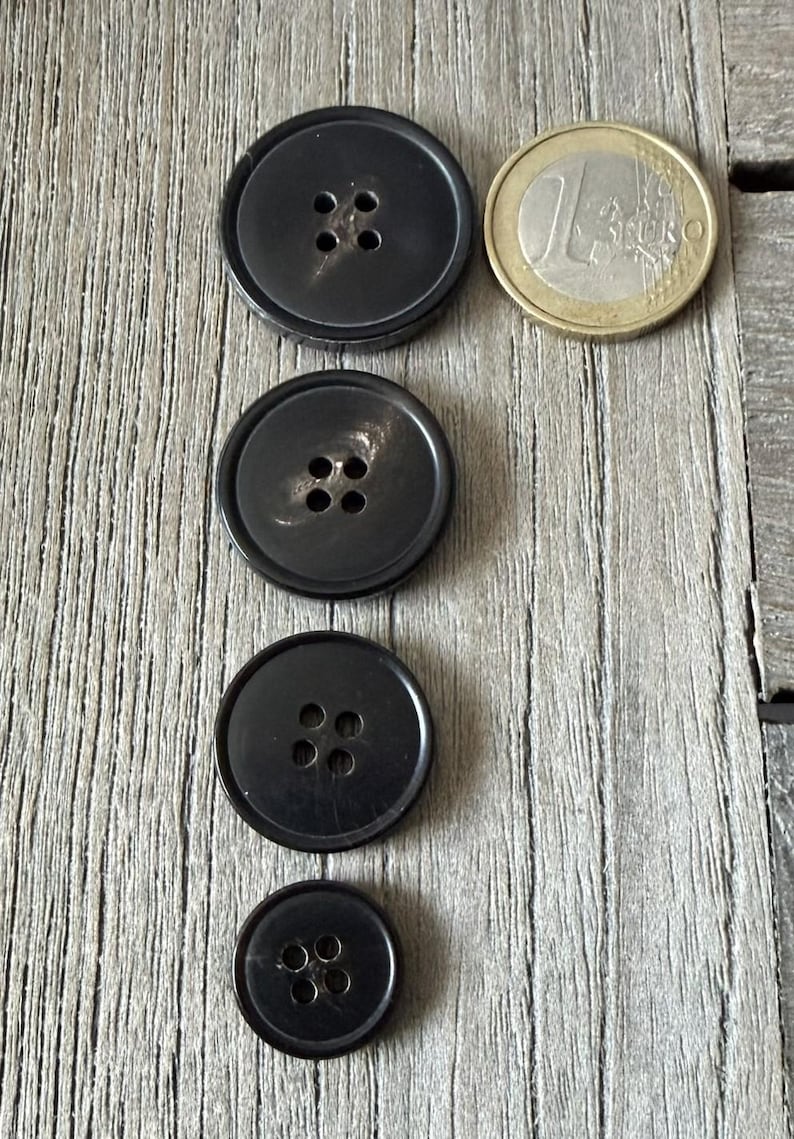 May include: Four black buttons of varying sizes with four holes each, arranged vertically on a wooden surface. A one Euro coin is next to the largest button. The buttons are round and have a glossy finish.