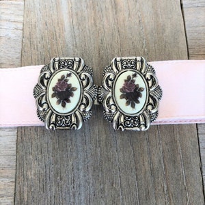 May include: A pair of silver-tone belt buckles with a floral design. The buckles have a white background with a pink rose and green leaves.