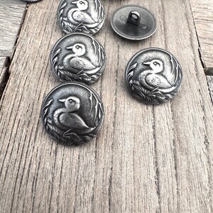 May include: A collection of silver metal buttons with a duck design. The buttons are round and have a raised design of a duck in a pond.