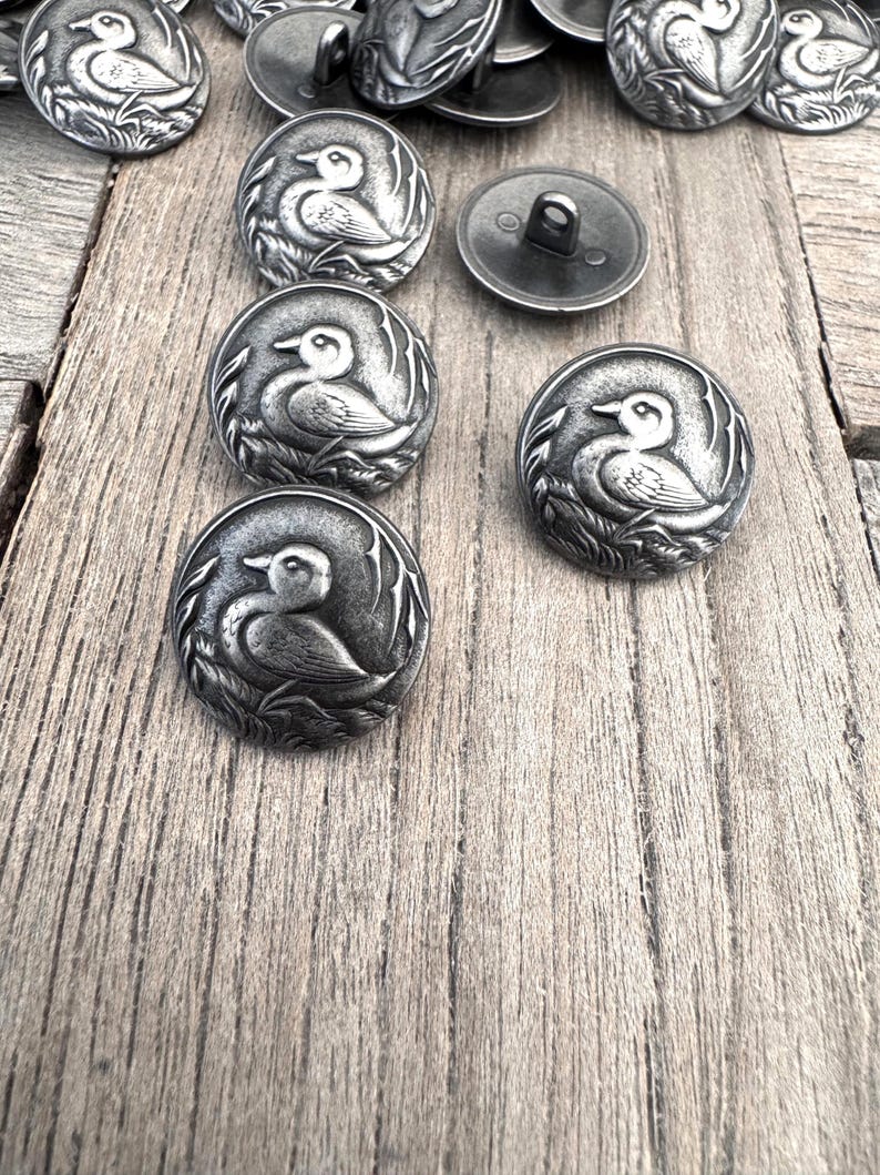 May include: A collection of silver metal buttons with a duck design. The buttons are round and have a raised design of a duck in a pond.