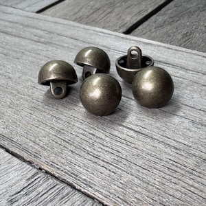 May include: Five antique bronze buttons are displayed. The buttons are dome-shaped with a loop on the underside for sewing. They are arranged on a weathered, grey wooden surface. Each button is approximately 1.5 cm in diameter.
