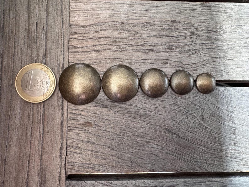 Peut inclure: Une rang&eacute;e de six clous ronds en m&eacute;tal dor&eacute; de tailles diff&eacute;rentes, dispos&eacute;s du plus grand au plus petit, sur une surface en bois. Une pi&egrave;ce d'un euro se trouve &agrave; gauche des clous.