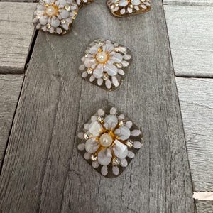 May include: A collection of decorative floral embellishments. Each piece features a white flower design with a central pearl and surrounding clear and gold accents. The embellishments are arranged on a gray wooden surface.