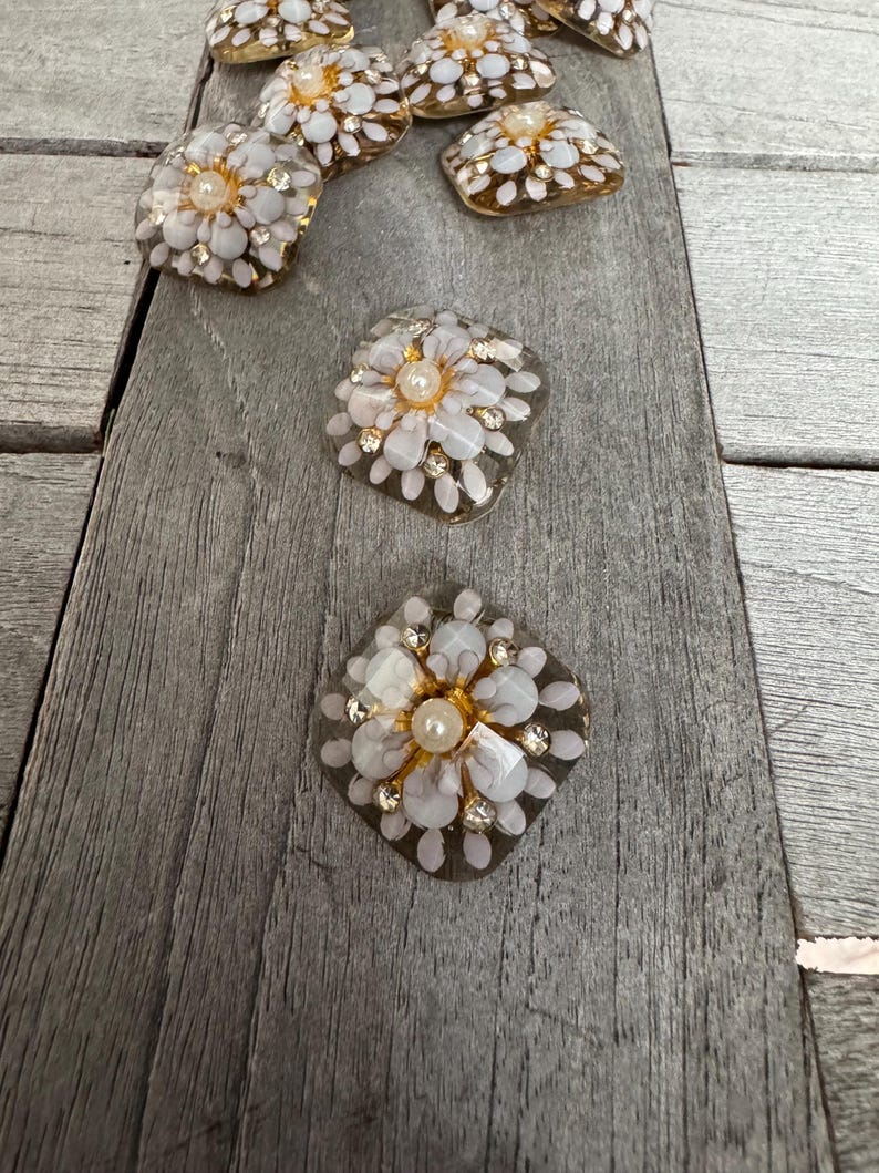 May include: A collection of decorative floral embellishments. Each piece features a white flower design with a central pearl and surrounding clear and gold accents. The embellishments are arranged on a gray wooden surface.