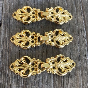May include: Three gold-toned metal decorative clasps with an ornate design. Each clasp has two pieces that connect together.