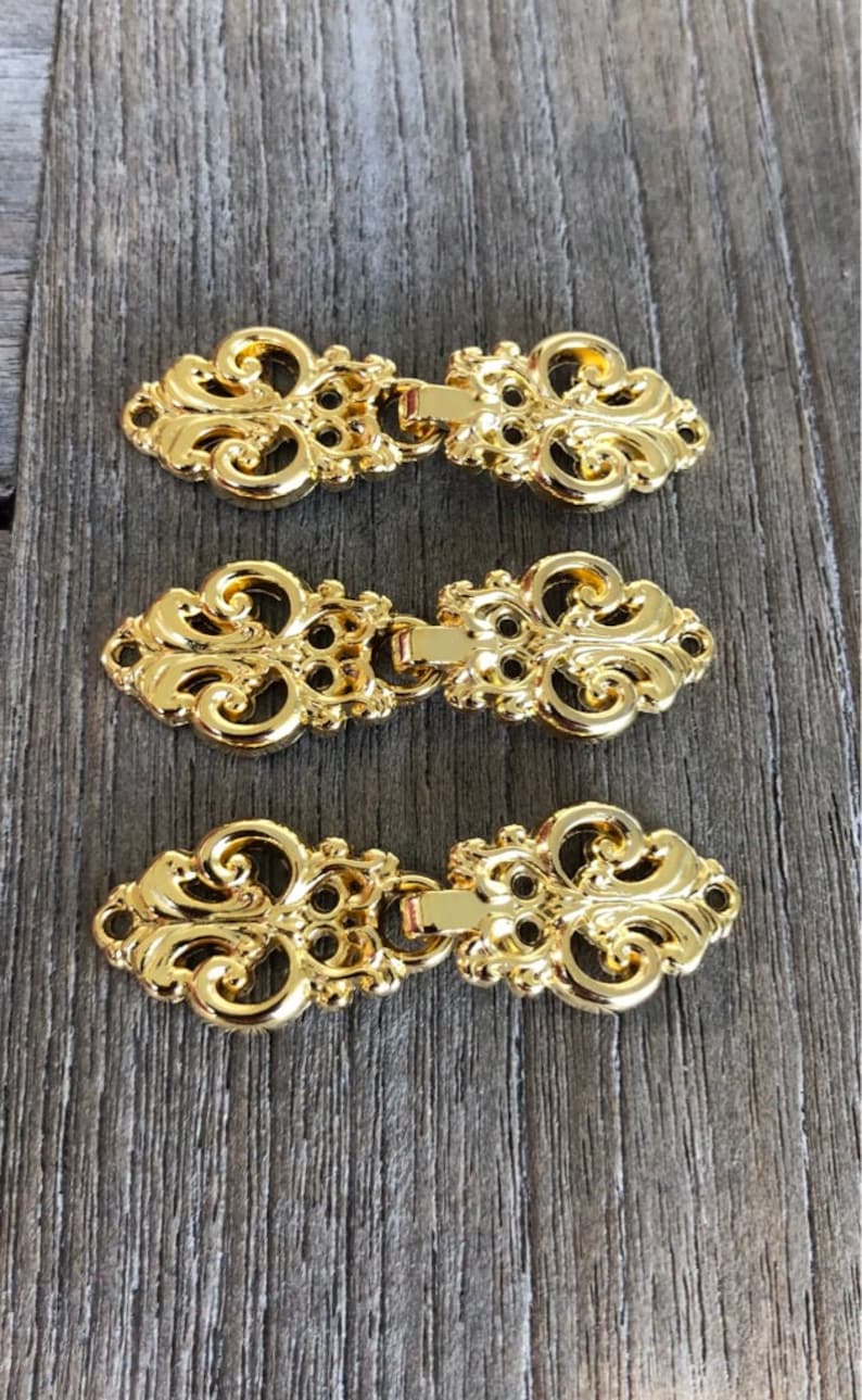 May include: Three gold-toned metal decorative clasps with an ornate design. Each clasp has two pieces that connect together.