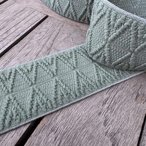 May include: A roll of sage green woven ribbon with a diamond pattern. The ribbon is displayed on a weathered wooden surface, highlighting its texture and design. Suitable for craft projects, the ribbon is a versatile material.