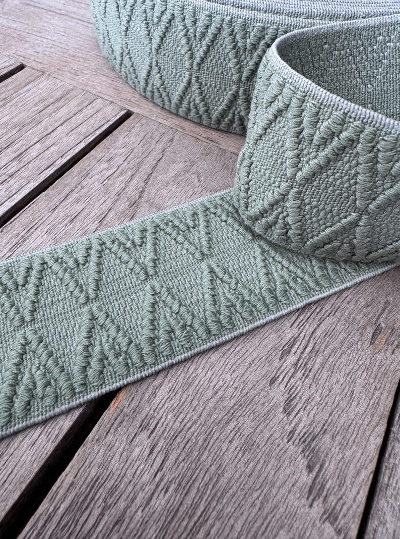 May include: A roll of sage green woven ribbon with a diamond pattern. The ribbon is displayed on a weathered wooden surface, highlighting its texture and design. Suitable for craft projects, the ribbon is a versatile material.