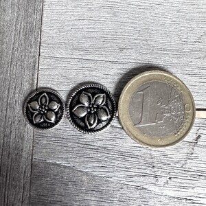 Może przedstawiać: Dwa srebrne metalowe guziki z kwiatowym wzorem. Guziki są r&oacute;żnych rozmiar&oacute;w i pokazane są obok monety euro w celu por&oacute;wnania.