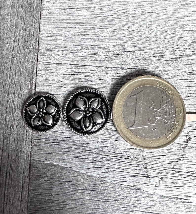 Może przedstawiać: Dwa srebrne metalowe guziki z kwiatowym wzorem. Guziki są r&oacute;żnych rozmiar&oacute;w i pokazane są obok monety euro w celu por&oacute;wnania.