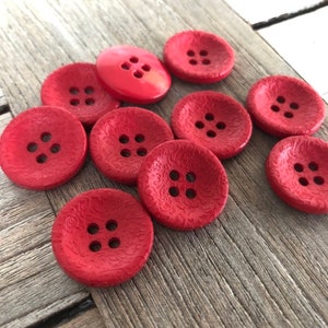 May include: A collection of ten red buttons with four holes each. The buttons have a textured surface and are arranged on a wooden surface.