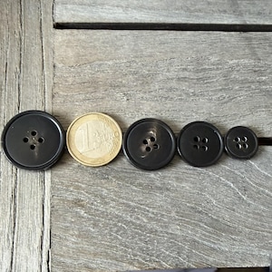 May include: A collection of dark gray buttons of varying sizes, arranged next to a one Euro coin. The buttons have four holes each and are made of a smooth, possibly plastic material. The buttons are on a weathered wooden surface.