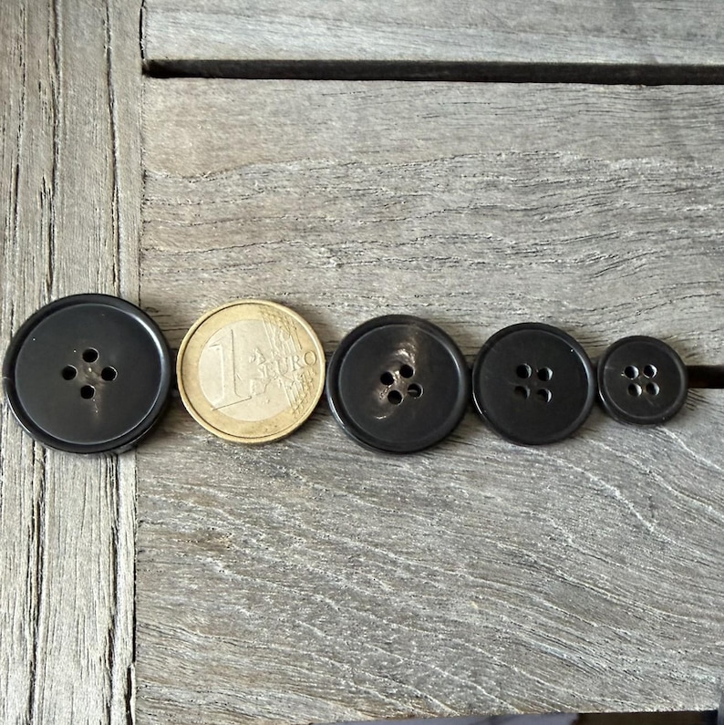May include: A collection of dark gray buttons of varying sizes, arranged next to a one Euro coin. The buttons have four holes each and are made of a smooth, possibly plastic material. The buttons are on a weathered wooden surface.