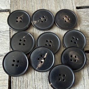 May include: A collection of ten large, round, black buttons with four holes each. The buttons are made of a dark material with a subtle, natural grain. They are arranged on a weathered wooden surface.