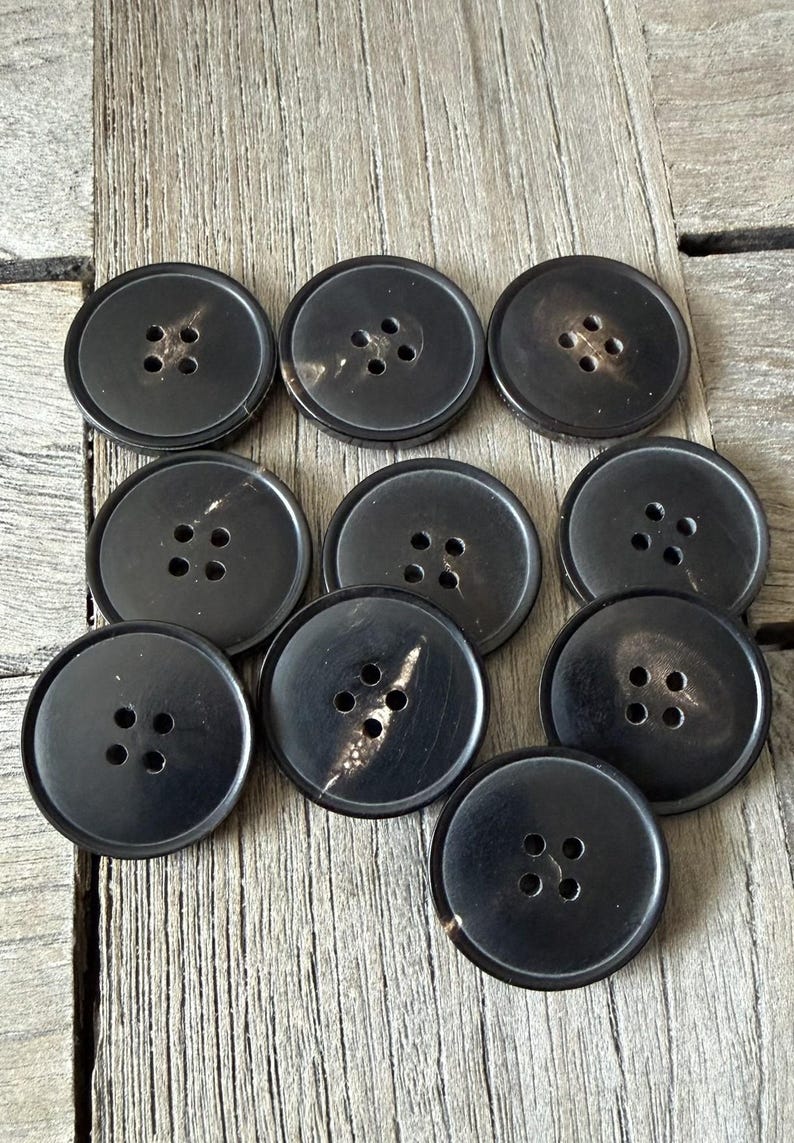 May include: A collection of ten large, round, black buttons with four holes each. The buttons are made of a dark material with a subtle, natural grain. They are arranged on a weathered wooden surface.