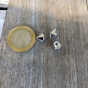 K&ouml;nnte beinhalten: Drei silberne Metallkn&ouml;pfe in Kegelform mit einem Loch oben. Die Kn&ouml;pfe liegen auf einer Holzoberfl&auml;che.