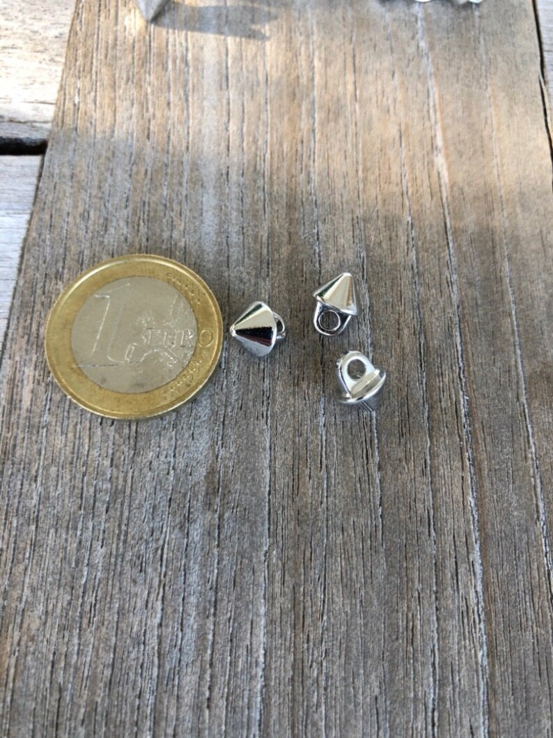 K&ouml;nnte beinhalten: Drei silberne Metallkn&ouml;pfe in Kegelform mit einem Loch oben. Die Kn&ouml;pfe liegen auf einer Holzoberfl&auml;che.